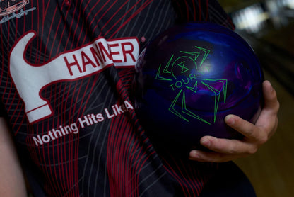 Person holding a blue bowling ball with a logo, wearing a shirt with 'Hammer' branding.